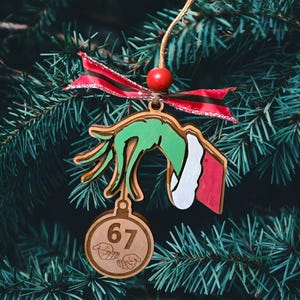 May include: Wooden Christmas tree ornament depicting a green hand with white and red details, holding a round ornament with the number 67. It's adorned with a red and black ribbon and a red bead, hanging on a fir tree.