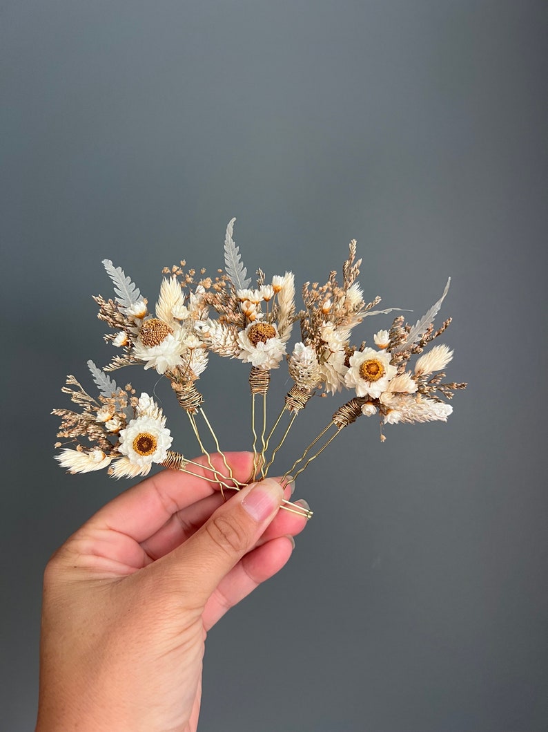 May include: Set of five gold hair pins decorated with dried flowers. The flowers are white and brown and include small daisies and dried grasses.