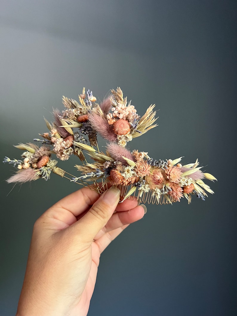 May include: A gold hair comb adorned with dried flowers in shades of pink, beige, and lavender. The comb features a variety of dried flowers, including small pink roses, fluffy white flowers, and dried grasses.