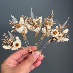 May include: Set of five gold hair pins decorated with dried flowers. The flowers are white and beige, with a variety of textures and shapes.