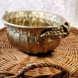 May include: A gold-toned metal bowl with a decorative grape design on the handles. The bowl is sitting on a woven wicker surface.