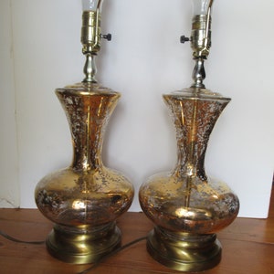 Peut inclure: Deux lampes de table vintage avec une finition dorée et argentée mouchetée. Les lampes ont une base bulbeuse et un col fin, avec des accents métalliques couleur laiton et une ampoule en haut. Les lampes mesurent environ 51 cm de haut.
