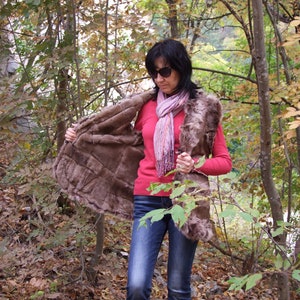 May include: A brown fur vest with a quilted design is open, showing the lining. The person in the image is wearing a red long-sleeved top, blue jeans, and a pink scarf. The background is a forest with autumn leaves.
