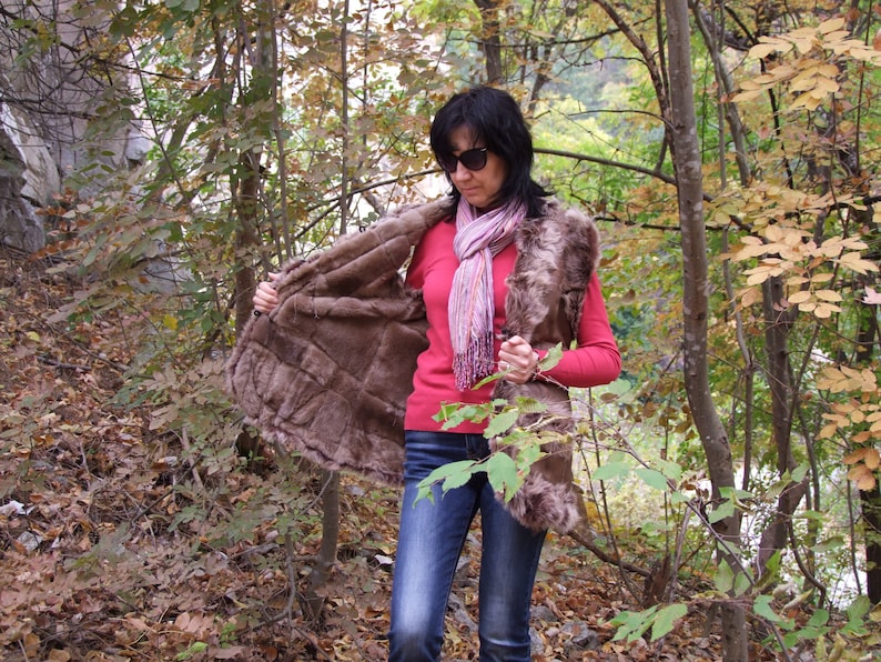 May include: A brown fur vest with a quilted design is open, showing the lining. The person in the image is wearing a red long-sleeved top, blue jeans, and a pink scarf. The background is a forest with autumn leaves.