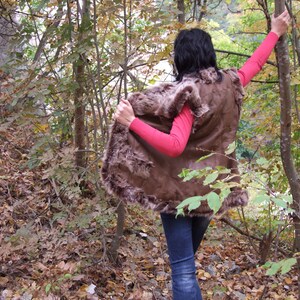 May include: A brown fur-trimmed vest is worn over a pink long-sleeved top and blue jeans. The person is in a forest with trees and fallen leaves.