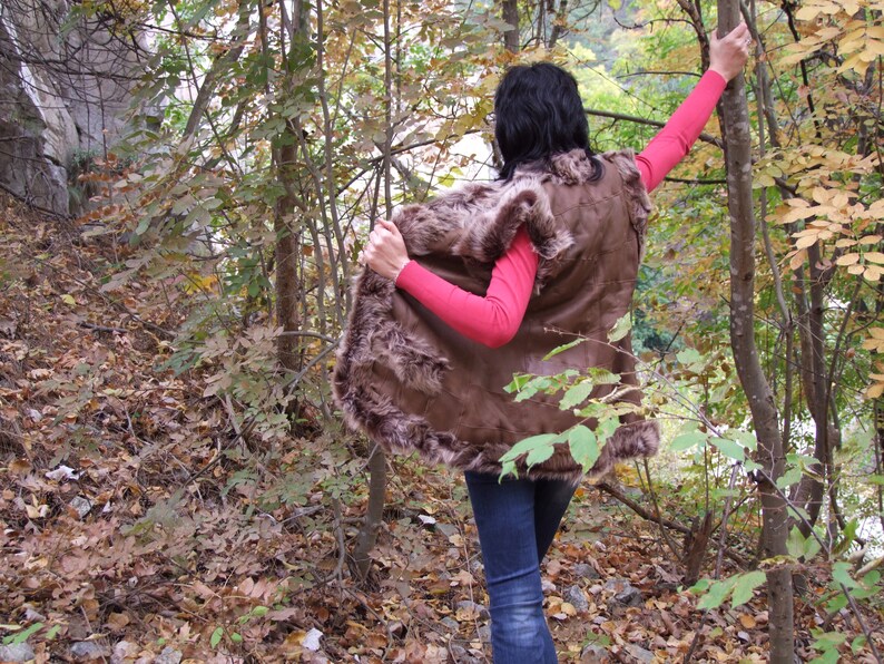 May include: A brown fur-trimmed vest is worn over a pink long-sleeved top and blue jeans. The person is in a forest with trees and fallen leaves.