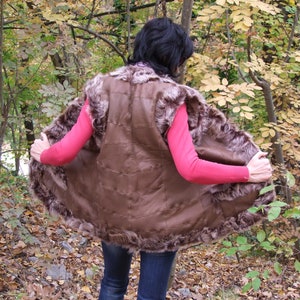 May include: A brown fur vest with a leather interior. The vest has a fur trim around the edges and a quilted design on the back. The person is wearing a pink long-sleeve top and blue jeans.