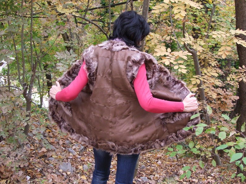 May include: A brown fur vest with a leather interior. The vest has a fur trim around the edges and a quilted design on the back. The person is wearing a pink long-sleeve top and blue jeans.