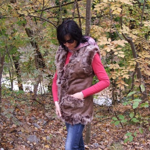 May include: A brown faux fur vest with a suede-like exterior. The vest has a pocket and is worn over a red long-sleeved shirt and blue jeans. The background features a forest with trees and autumn foliage.