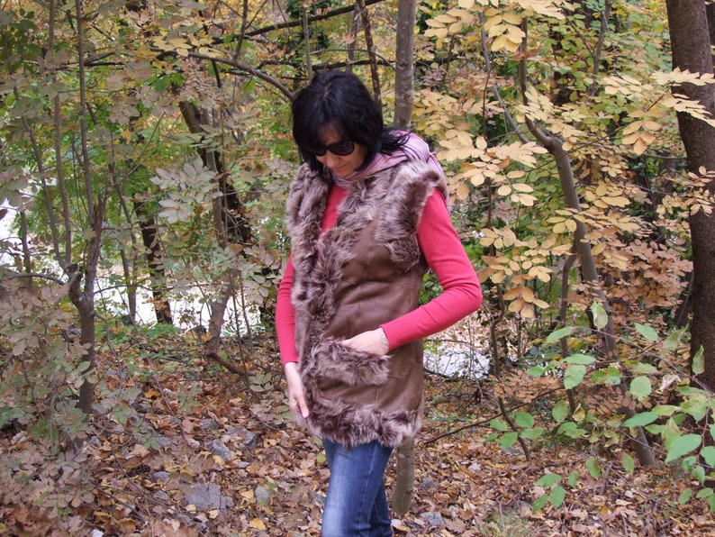 May include: A brown faux fur vest with a suede-like exterior. The vest has a pocket and is worn over a red long-sleeved shirt and blue jeans. The background features a forest with trees and autumn foliage.