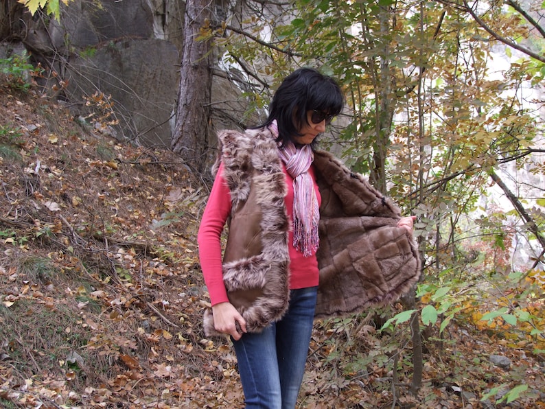 May include: A brown fur vest with a textured exterior and a quilted interior. The vest has two front pockets and a hood. The person is wearing a pink long-sleeve top, blue jeans, and a pink and white scarf.