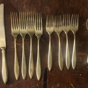 May include: A set of twelve pieces of silver-toned flatware arranged in three groups. The collection includes four dinner knives with smooth handles, four large forks with four tines each, and four spoons with oval bowls and slender handles.
