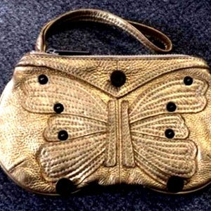 May include: A gold-colored handbag with a butterfly design. The bag features a handle and a zipper closure. The butterfly wings are detailed with stitching and adorned with black circular accents.