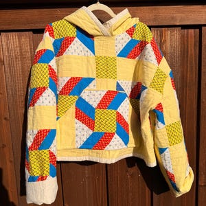 May include: A hooded pullover sweatshirt constructed from quilted fabric featuring a patchwork pattern of yellow, red, and blue geometric shapes. The garment includes a front kangaroo pocket and long sleeves, with a cream-colored hood lining.