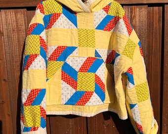 Puede incluir: Una sudadera con capucha confeccionada en tela acolchada con un patrón de retazos de formas geométricas amarillas, rojas y azules. La prenda incluye un bolsillo tipo canguro frontal, mangas largas y un forro de capucha color crema.