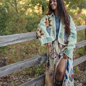 May include: A quilted jacket featuring a light green and white checkered pattern with sunflower motifs, worn over a blue denim button-down shirt and a long, multi-colored patchwork skirt with a high side slit, paired with dark brown leather boots.