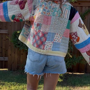May include: A quilted jacket constructed from various patchwork fabric squares in shades of pink, blue, white, and red. The garment features long sleeves with contrasting striped and floral patterns, a collar, and a visible interior label at the neck.
