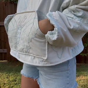 May include: A light blue and white quilted pullover jacket featuring a large front kangaroo pocket with an embroidered butterfly design. The jacket has long sleeves with elasticized cuffs and a high collar, constructed from a patchwork fabric.