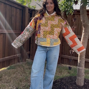 May include: A long-sleeved hooded pullover constructed from various quilted fabric patches in shades of orange, yellow, white, and brown. The garment features a mix of floral, plaid, and geometric patterns with visible stitching throughout the fabric.