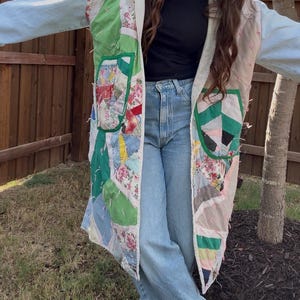 May include: A long, open-front coat constructed from repurposed patchwork quilt fabric featuring various floral and geometric patterns in shades of green, pink, and white, with light blue denim sleeves and two large patch pockets on the front.