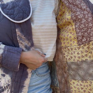 May include: A quilted jacket featuring a dark gray tie-dye exterior with white accents and a contrasting interior lining composed of various patchwork fabric squares in shades of yellow, brown, and white with floral and geometric patterns.