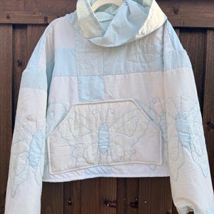 May include: A long-sleeve hooded sweatshirt constructed from a white and light blue patchwork quilt. The front features a large kangaroo pocket with a stitched butterfly design, while additional butterfly motifs are visible on the sleeves.