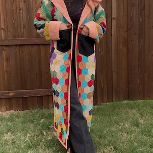 May include: A long, hooded coat featuring a multicolored patchwork design of small hexagons in shades of red, blue, yellow, green, and brown. The coat has a solid peach-colored trim along the front opening and hood, with two large black patch pockets on the front.