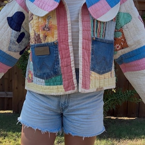 May include: A quilted patchwork jacket featuring a mix of floral, striped, and solid fabric panels in pink, blue, and cream. The jacket includes two dark blue denim pockets with the Wrangler logo patch, a large rounded collar, and a front opening.