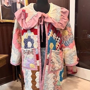 May include: A long-sleeved quilted jacket constructed from a patchwork of multicolored hexagonal fabric pieces. The jacket features a large, solid pink ruffled collar with a tie closure at the neck and two square patch pockets on the front.
