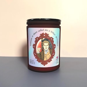 May include: A red glass candle jar with a black lid. The label features a portrait in a decorative frame and text that reads "Someone in there called me a Volturi, #FTF2025". The candle is labeled "Velvet Bite" and "Scent of a Killer".