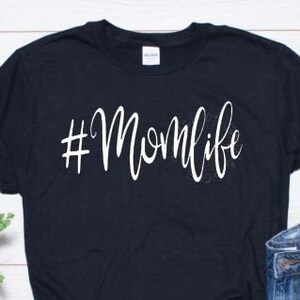May include: Black t-shirt with white text that says "#Momlife". The shirt is laying flat on a white wooden surface with a pair of blue denim shorts, a pair of white sneakers, and a pair of sunglasses.