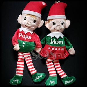 May include: Two plush Christmas elves with red hats, green and red outfits, and white striped stockings. The elves are personalized with the names "Pops" and "Mom".