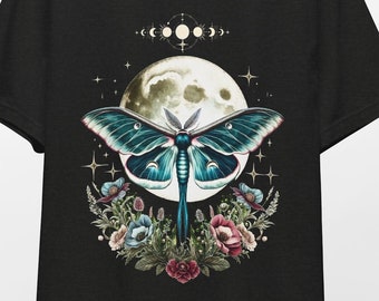 May include: Black t-shirt featuring a large teal moth with detailed wings, set against a full moon and floral arrangement. The design includes phases of the moon and stars, creating a celestial theme. The shirt is hanging on a wooden hanger.