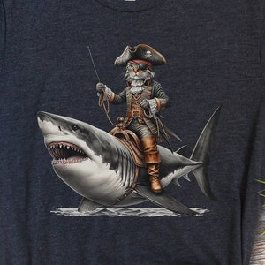 以下が含まれることがあります： 暗いグレーのTシャツに、白いサメに乗った猫の海賊のグラフィックがプリントされています。猫は海賊の帽子をかぶり、剣を持っています。