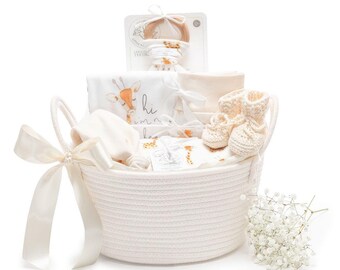 May include: A white woven rope basket containing baby items including a wooden teething ring, a white onesie with a giraffe print and the text hi i'm new here, a pair of cream-colored knit booties, a plain cream fabric item, and a white satin ribbon.