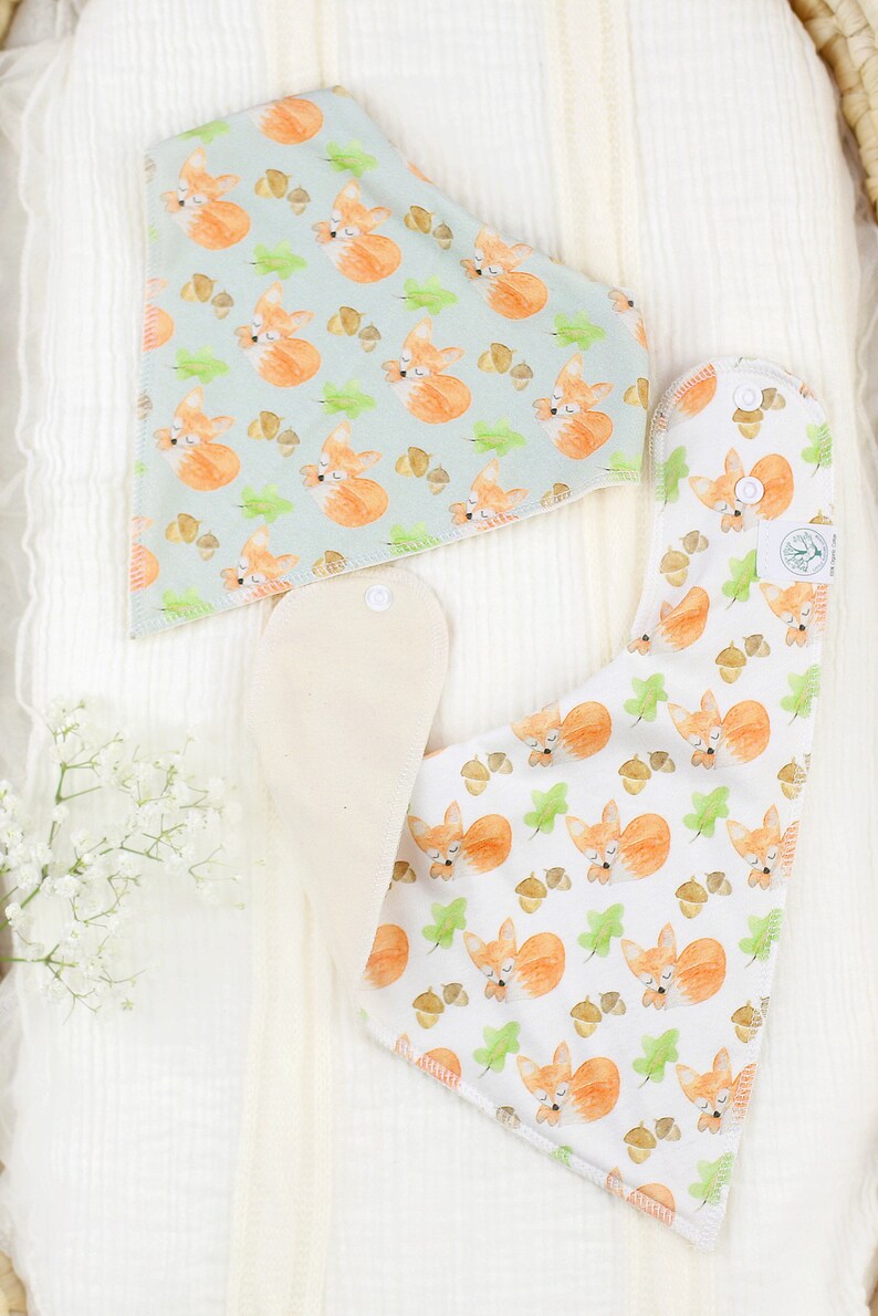 May include: Two baby bibs with a fox and acorn pattern. One bib is light blue with white foxes and the other is white with orange foxes. Both bibs have a white absorbent layer.