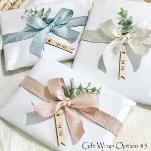 May include: Three white gift boxes wrapped with different colored ribbons and greenery. Each box has a wooden tag that says "For You". The text below the boxes reads "Gift Wrap Option $5 During checkout the box that says "This order is a gift""