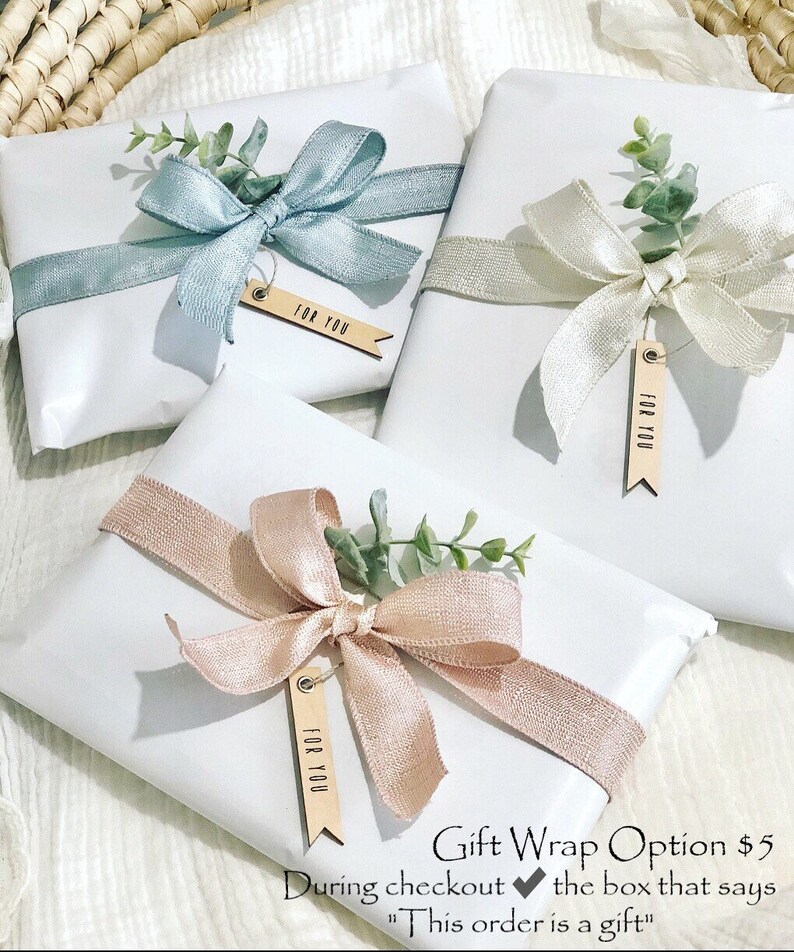 May include: Three white gift boxes wrapped with different colored ribbons and greenery. Each box has a wooden tag that says "For You". The text below the boxes reads "Gift Wrap Option $5 During checkout the box that says "This order is a gift""