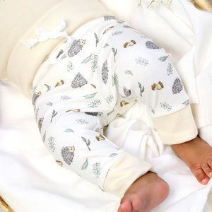 May include: White baby pants with a hedgehog and leaf pattern. The pants have a cream colored waistband and cuffs.