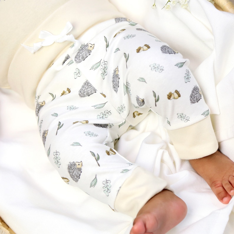 May include: White baby pants with a hedgehog and leaf pattern. The pants have a cream colored waistband and cuffs.