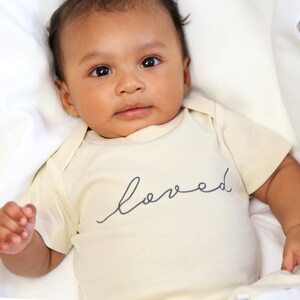 May include: A baby wearing a cream-colored onesie with the word "loved" written in black script on the front.