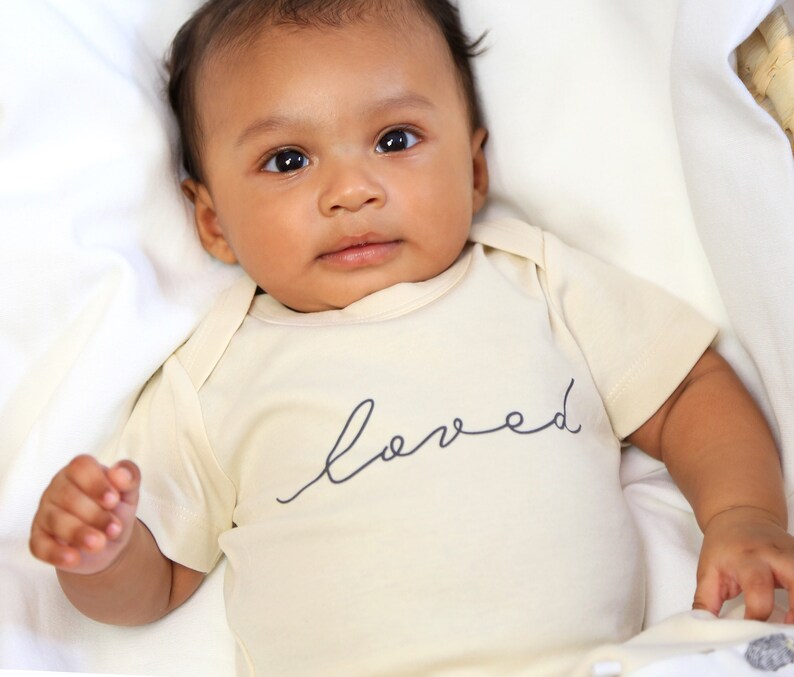 May include: A baby wearing a cream-colored onesie with the word "loved" written in black script on the front.