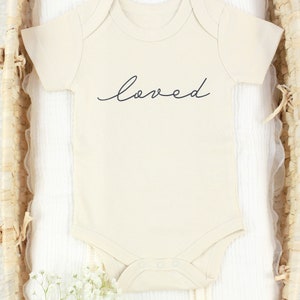 May include: A cream-colored baby onesie with short sleeves and the word "loved" written in black cursive script.