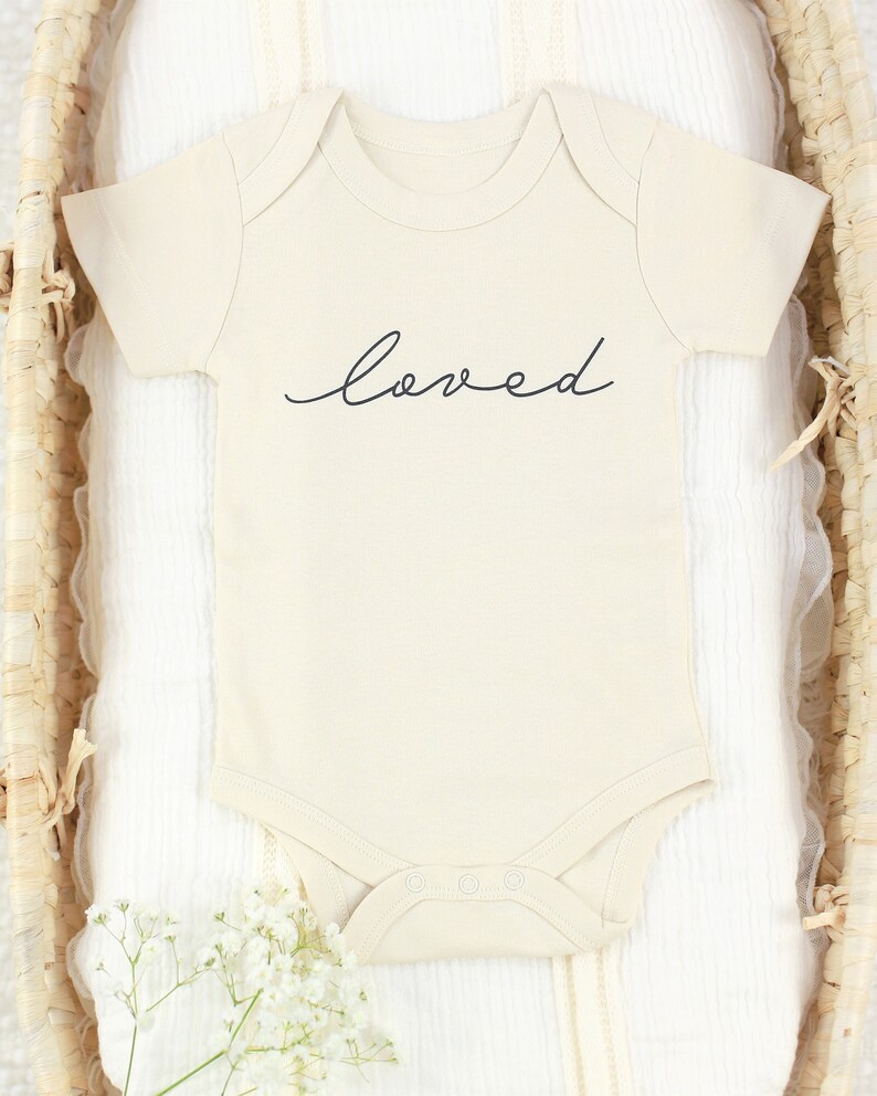 May include: A cream-colored baby onesie with short sleeves and the word "loved" written in black cursive script.