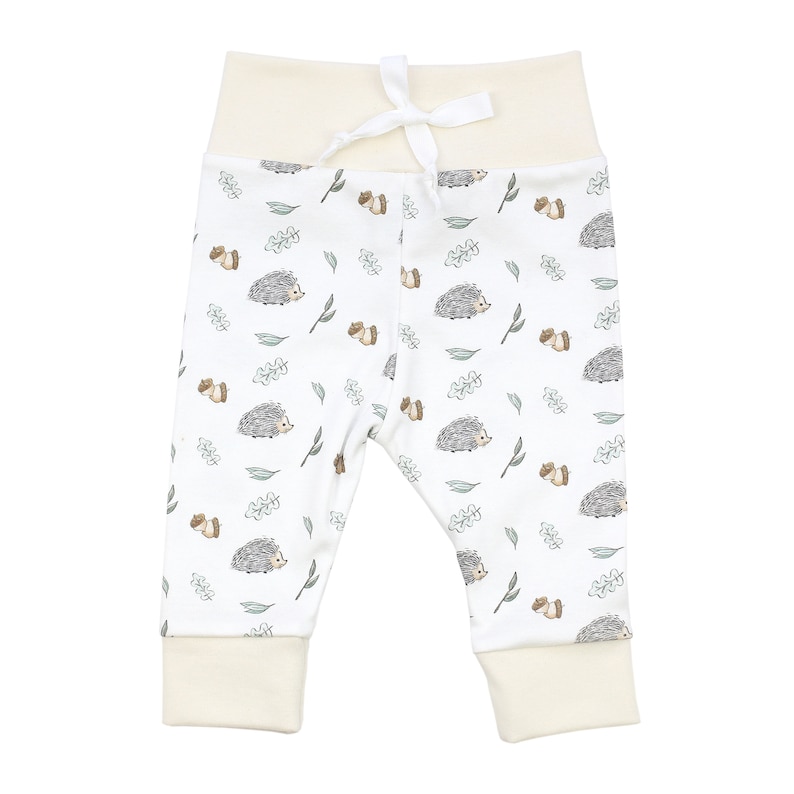 May include: White baby leggings with a cute hedgehog and leaf pattern. The leggings have a cream colored waistband and cuffs.