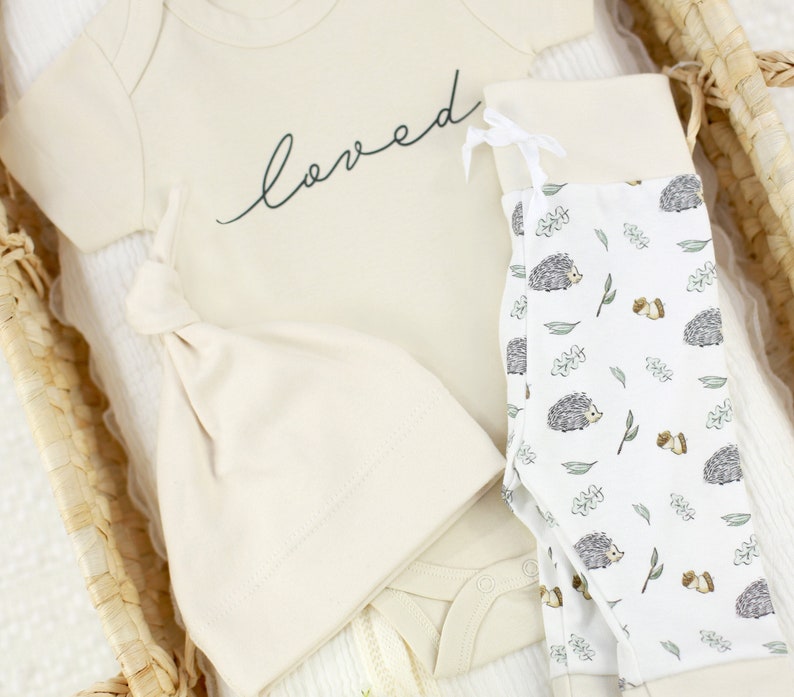 May include: A beige baby outfit set with a bodysuit, hat, and leggings. The bodysuit has the word "loved" written on it in black script. The leggings have a white background with a repeating pattern of hedgehogs, leaves, and acorns.