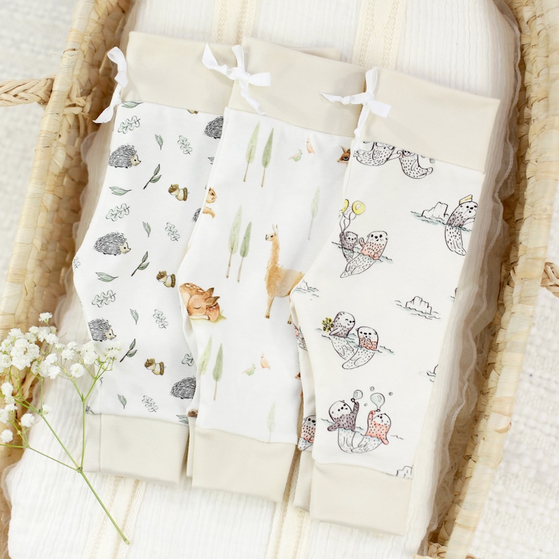 May include: Three pairs of baby leggings with different animal prints. The leggings are white with a cream colored waistband and have a variety of animal prints including hedgehogs, llamas, and otters.
