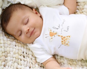 May include: A white baby onesie with a cute giraffe illustration and the text "hi i'm new here". The baby is sleeping on a white fuzzy blanket.