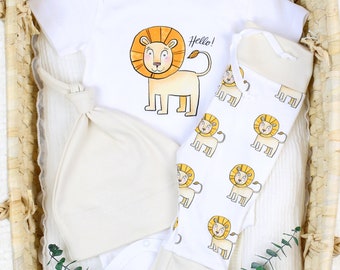May include: A white baby bodysuit, pants, and hat set. The bodysuit features a cartoon lion with the word "Hello!". The pants and hat are cream-colored. The pants have a repeating lion pattern. The set is displayed in a woven basket.