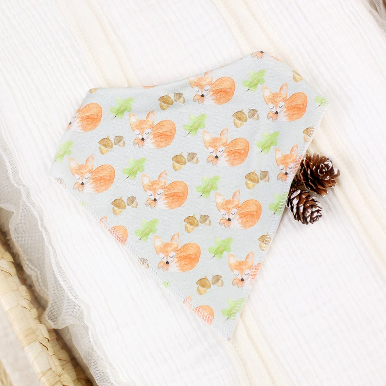 May include: A light gray triangular bandana with a repeating pattern of sleeping foxes and acorns. The foxes are orange with white accents and the acorns are brown.
